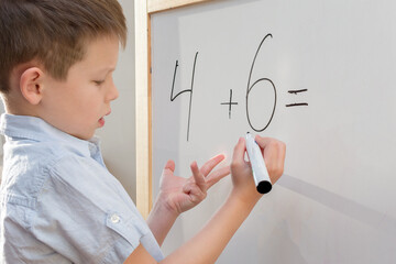 A preschool-age boy with a funny facial expression stands at a white board and decides an example in mathematics. The concept is to prepare the child for school, to obtain basic knowledge and skills.