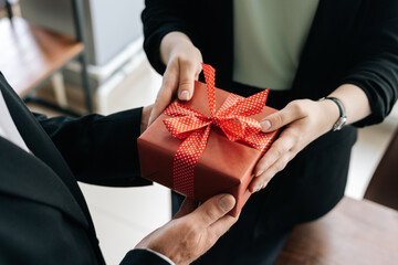 Close-up view of hands of unrecognizable woman giving red gift box tied to bow handed to man. Giving gifts during the Christmas, Happy New Year and Happy Birthday at office.