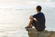 Thoughtful Young Man Sitting Alone On Top Of A Cliff Above The Ocean View At Sunset.