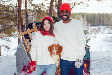 Wall Mural - Multiracial couple are sitting in the trunk of a car with a dog. A couple of lovers came to the forest for a Christmas tree.