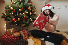 Merry Christmas! Happy Woman In Santa Hat Holding Christmas Gift  At Christmas Tree With Lights