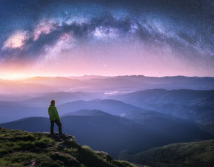 Wall Mural - man on the mountain peak and arched milky way over mountains in low clouds at night. landscape with 