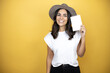 Beautiful woman wearing casual white t-shirt and a hat standing over yellow background smiling and showing blank notebook in her hand