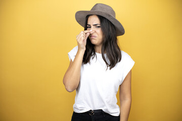 Wall Mural - Beautiful woman wearing casual white t-shirt and a hat standing over yellow background smelling something stinky and disgusting, intolerable smell, holding breath with fingers on nose