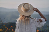 Fototapeta Koty - The blonde in a linen dress and straw hat walks among herbs and wildflowers on a meadow in the mountains. A woman in the Boho style, resting in the countryside, a simple village lifestyle