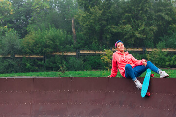 Portrait of a trendy pretty young gir with short colored hair sitting next to the skateboard court with her plastic skateboard.