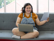 © Bangkok Click Studio - fat asian businesswoman wearing casual cloth with headphones smiling and look forward to a camera holding a paper in her hand with a laptop computer on her lap sitting on a couch working at home