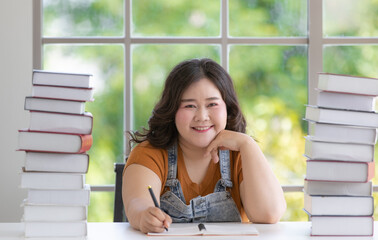 Wall Mural - Chubby asian woman student feel happy sitting smiling and holding pen writing on a book on a table with a stack of books in a room of the house. Study and work from home concept. 