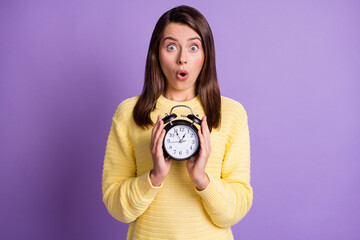 Wall Mural - Photo portrait of upset surprised woman holding black alarm clock with two hands isolated on vivid purple colored background