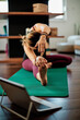 © chika_milan - Middle-aged flexible woman sitting at home in head to knee yoga posture and following instruction over laptop.