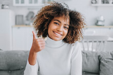 Happy Young Woman With Trendy Kinky Hairstyle And Dark Skin Smiles Showing Thumbs-up Against Modern White Kitchen Close View