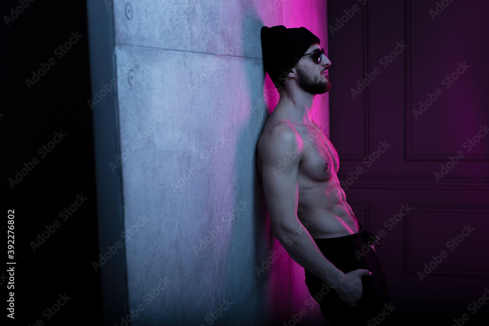 Portrait of a shirtless fit man in hat posing in dark studio with concrete wall with purple neon complementary color. - obrazy, fototapety, plakaty 