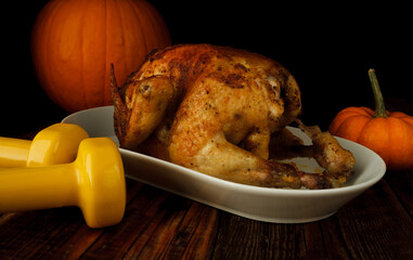 Roasted homemade chicken for Thanksgiving dinner, with pumpkins and two dumbbells. Autumn composition on wooden table. Healthy fitness lifestyle concept, cheat day temptation vs sticking to the diet.