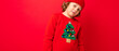 © Ivan Traimak - cool teenager in a red Christmas sweater fooling around against the background of a red wall, a warm hat and a sweater with a Christmas tree