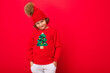 © Ivan Traimak - cool blond kid in warm hat and sweater with christmas tree on red background fooling around, christmas concept