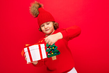 a cool boy with curls on a red wall background in a sweater with a Christmas tree holding a gift box in his hands