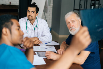 Wall Mural - African medical man doctor making report to colleagues on results of patient chest X-ray image in office room on background of window with X-ray and MRI scans. Concept of medicine and health care.