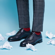 A Man Stands In Elegant Black Shoes On A Blue Background Among Crumpled Notes Of Paper.