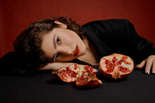 A Beautiful Young Woman In A Black Jacket Is Sitting At A Table On A Red Background. Holds Halves And Pieces Of Pomegranate Fruit