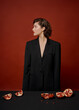 © Tatiana Timofeeva/Stocksy - a beautiful young woman in a black jacket is sitting at a table on a red background. holds halves and pieces of pomegranate fruit