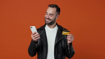 Wall Mural - Smiling laughing young bearded man in black leather jacket isolated on orange background studio. People lifestyle concept. Using mobile call phone hold credit bank card say yes doing winner gesture