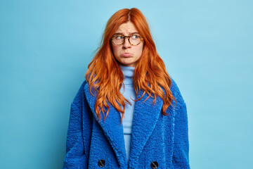 Wall Mural - Young displeased foxy woman purses lips and looks with sullen expression aside dressed in turtleneck and blue winter coat poses indoor expresses negative emotions. Stressed young ginger female