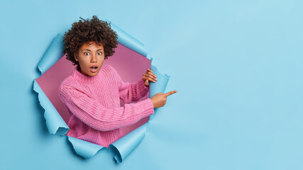 Poster - Shocked dark skinned woman with curly hair points away on blank space sees something incredible and breathtaking wears casual knitted sweater breaks through paper background. Omg look there.