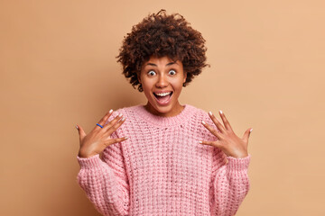 Wall Mural - Excited cheerful dark skinned woman raises palms and smiles broadly has surprised face expression wears casual knitted jumper expresses positive emotions poses against brown studio background