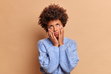 Wall Mural - Bored frustrated ethnic woman touches face has fatigue expression being sick and tired of weekdays needs rest dressed in blue sweater isolated over brown background. Negative feelings concept