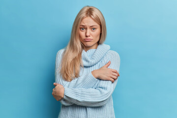 Wall Mural - Young dissatisfied blonde woman embraces herself looks with sad expression being hurt dressed in casual jumper isolated over blue background. Frustrated miserble upset female model poses indoor