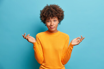 Wall Mural - Hesitant dark skinned woman has puzzled expression shrugs shoulders looks confused dressed in casual clothes isolated over blue background. Indecisive Afro American female makes doubt gesture