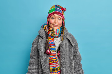 Wall Mural - Horizontal shot of surprised young Asian woman with rosy cheeks wears knitted hat scarf around neck and warm fur coat dresses for cold weather isolated over blue background. Winter time concept