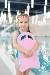 Wall Mural - Cute little girl in swimwear and goggles holding pink plastic swimming board