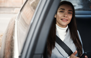 Elegant businesswoman sitting on backseat of her car with seatbelt, using mobile phone. Smiling woman texting message while driving to office