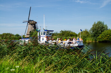 Wall Mural - Kinderdijk, Netherlands, August 2019. A historic windmill, in perfect condition, in the Dutch countryside crisscrossed by canals lined with tall green grasses. A tourist boat is passing.