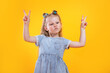 Funny little girl showing peace gesture on yellow background..