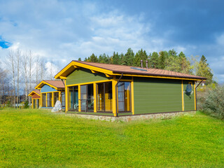 Wall Mural - Country cottages in a minimalistic style. Wooden cottages on a blue sky background. Forest behind cottage community. Several one-story houses stand side by side. Lawn in front of village houses.