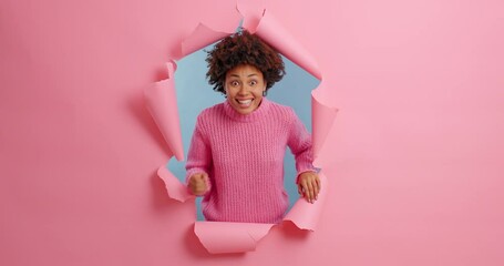 Wall Mural - Beautiful dark skinned woman makes winning gesture exclaims yes feels very glad celebrates success dressed in casual clothing poses through torn hole of pink background. Celebration concept.