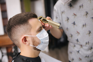 Professional hairdresser uses a hair clipper for fringing hair for a handsome bearder man. Client with face mask protect from the coronavirus. The hairdresser also wears a mask