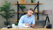© viacheslav - Serious confident gray-haired business man manager CEO sitting on chair at table in home office wears wireless earphones listening music writes information to notebook. Man doing English homework