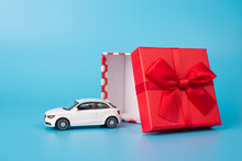 Giving And Receiving Gifts Concept. Close Up Photo Of White Toy Car Beside Open Red Giftbox With Bow Isolated On Blue Background
