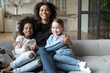 © fizkes - Portrait of smiling African American mother hugging two little daughters, multiracial family concept, sitting on cozy couch in living room, posing for photo, looking at camera, adopted child
