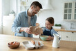 Smiling father and adorable little daughter cooking dough for pancakes in kitchen, standing at wooden table, happy dad holding flour, helping to cute girl child, family enjoying leisure time together
