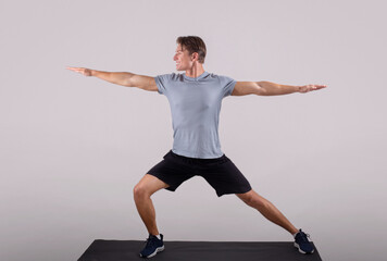 Sticker - Handsome young man in sportswear doing warrior yoga pose on sports mat over light background, full length portrait