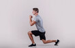 Side view of young fit guy in sportswear doing lunge on light studio background