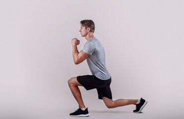 Wall Mural - Side view of young fit guy in sportswear doing lunge on light studio background