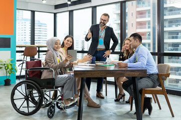 Asian Muslim business woman in hijab headscarf sitting on wheelchair presenting of her work to corporate colleagues in meeting in the modern office. diverse corporate colleagues and multicultural