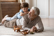 © fizkes - Overjoyed mature grandfather playing with adorable grandson at home, older man lying on warm floor with cute little boy grandchild on back, family spending leisure time together, having fun
