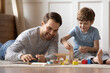 © fizkes - Close up happy loving father playing with adorable son at home, lying on warm floor, smiling dad and little boy kid constructing with wooden colorful blocks, enjoying leisure time in living room