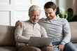© fizkes - Happy senior father with adult son using laptop at home together, sitting on cozy couch, hugging, smiling mature grandfather with grandson looking at computer screen, young man helping to senior dad